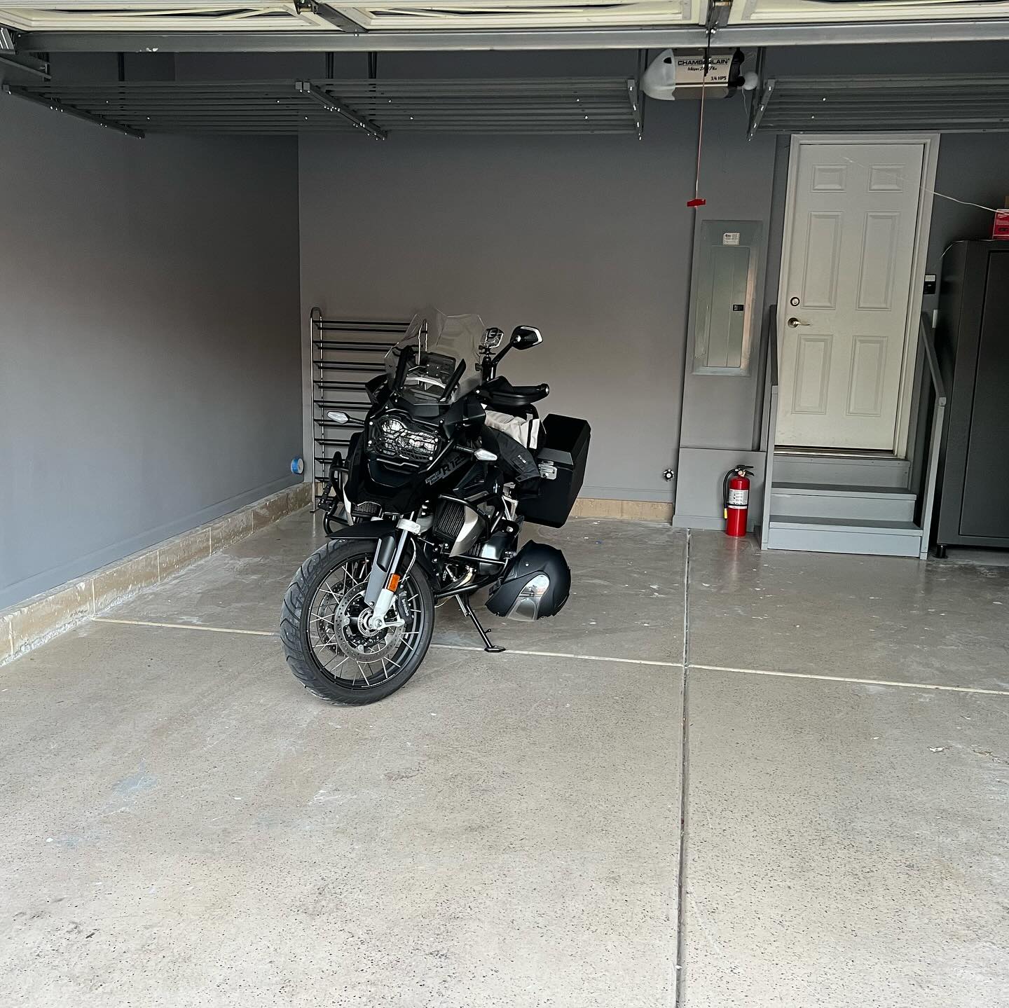 A motorcycle is parked inside a clean, spacious garage with gray walls, a fire extinguisher, and a closed white door leading outside.