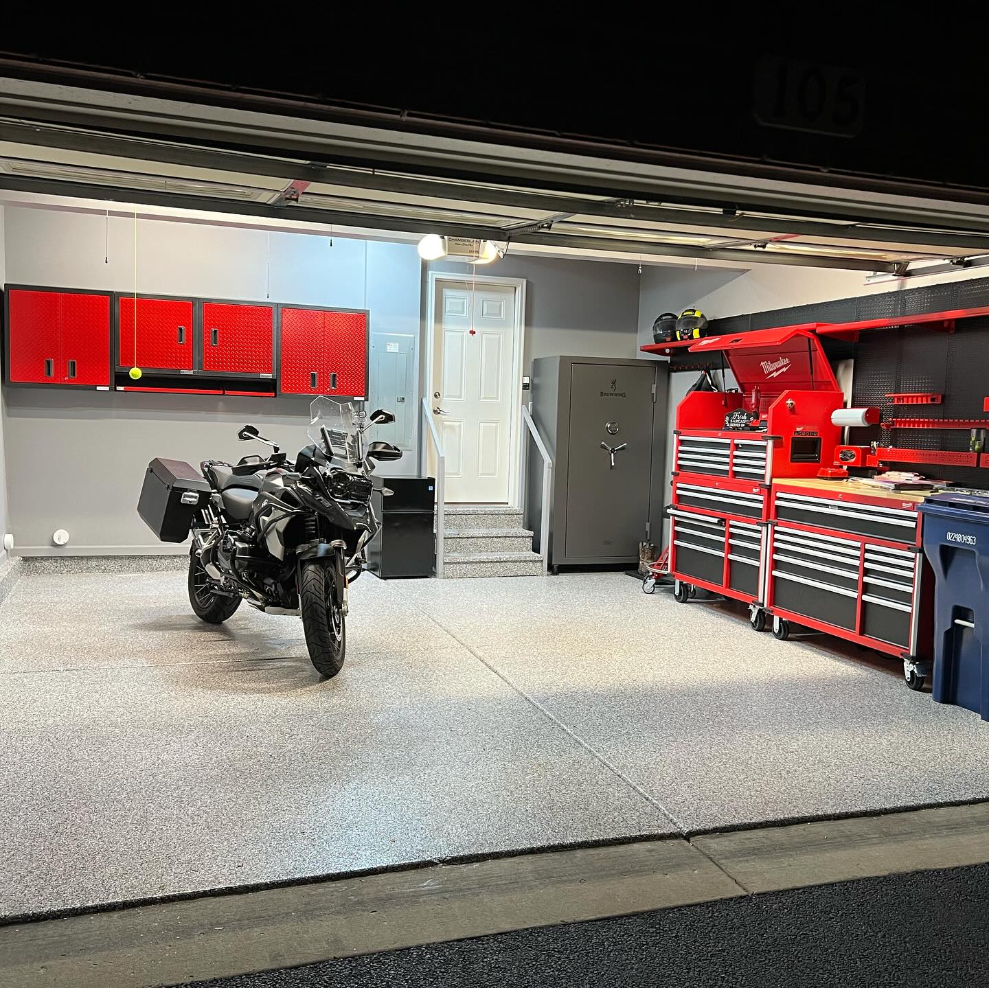 A well-organized garage with a black motorcycle, red tool cabinets, and a safe. Clean floor and walls, with helmets on a shelf.