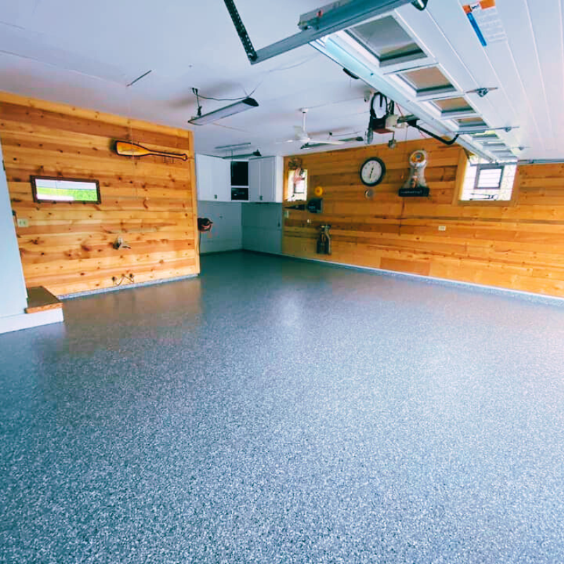 Spacious garage with speckled flooring, wooden walls, and ceiling fans. Includes a clock, cabinets, and a mounted paddle for decoration.
